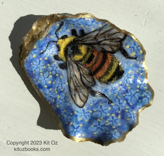 A small oyster shell with a yellow, orange, and black bumble bee painted on it. Background is a multitude of ceanothus blossoms in blue/lavender.
